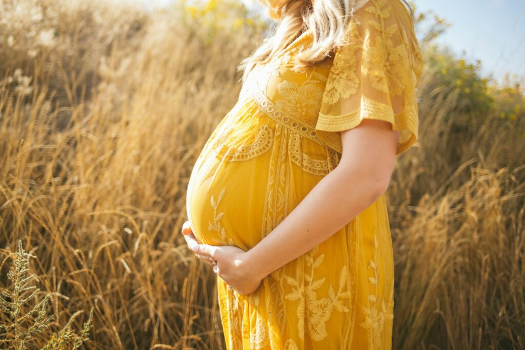 Pregnant woman wearing yellow floral dress standing in a field. Therapeutic Massage Treatment